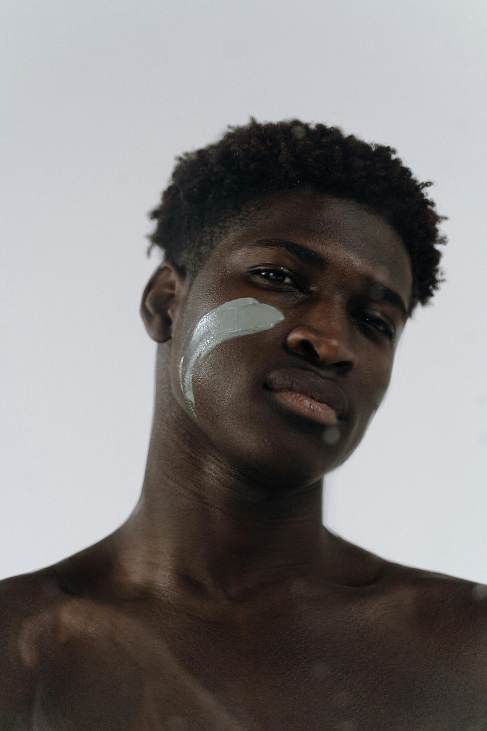 Close-up of a shirtless man with clay mask on cheek against white background.