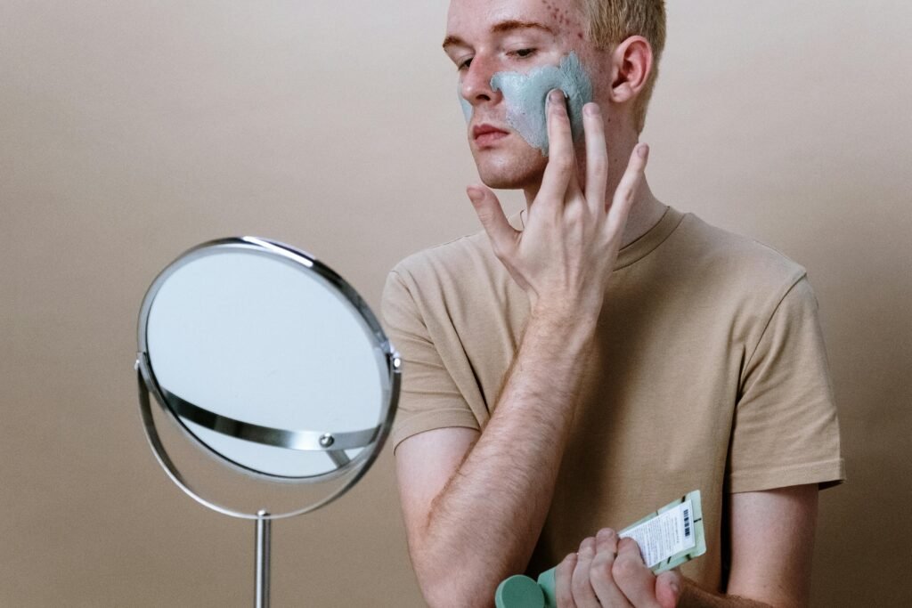 Young man applying clay mask for skincare routine indoors with a mirror.