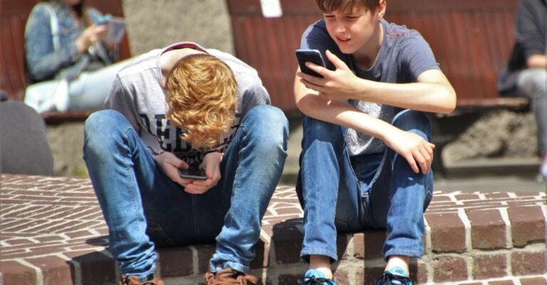Two teenagers seated outdoors, focused on their smartphones, embracing modern technology.